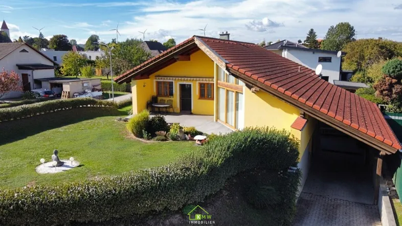 Gelbes Einfamilienhaus mit rotem Dach, großem Garten und Carport unter blauem Himmel.