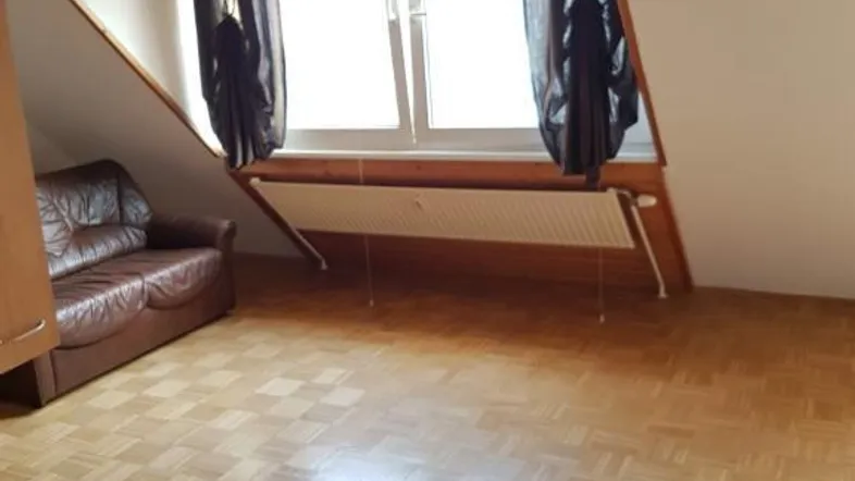 Helles Dachzimmer mit schrägen Wänden, Parkettboden und einem braunen Ledersofa.