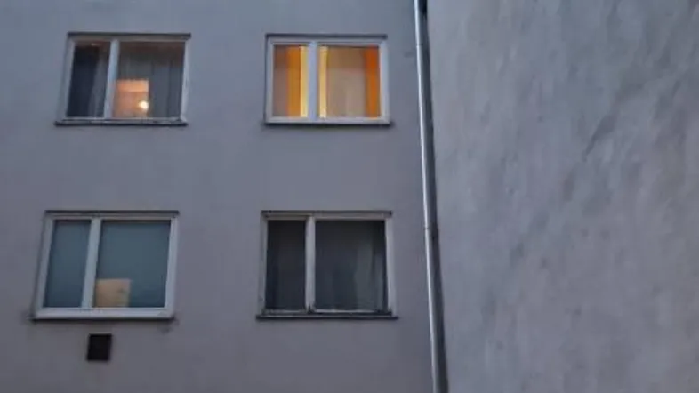 Fassade eines Mehrfamilienhauses bei Dämmerung mit beleuchteten Fenstern im oberen Stockwerk.