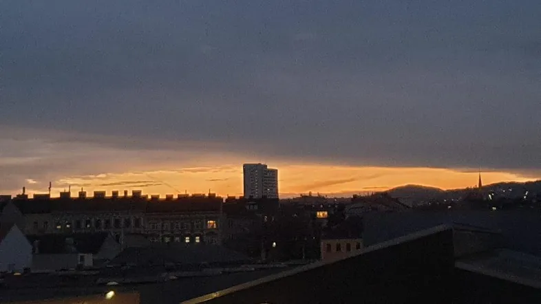Stadtansicht bei Sonnenuntergang mit orangefarbenem Himmel über den Dächern und einem fernen Hügel.