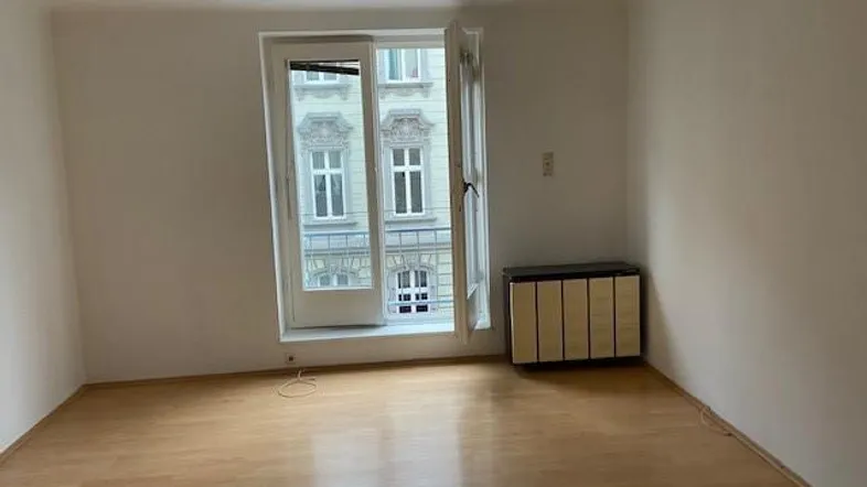 Helles, leeres Zimmer mit Parkettboden und großem Fenster, das auf ein gegenüberliegendes Gebäude blickt.
