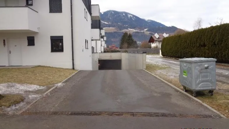 Außenansicht eines modernen Gebäudes mit Zufahrt zur Tiefgarage und Bergblick im Hintergrund.