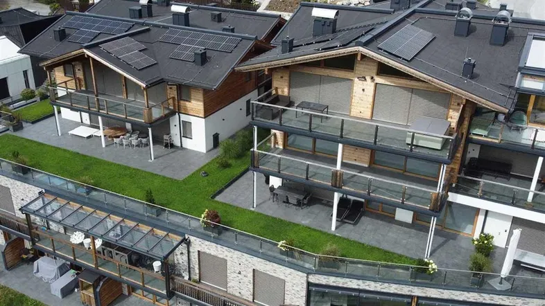 Luftaufnahme moderner Chalet-Gebäude mit Solarpaneelen, Balkonen und Terrassen in alpiner Umgebung.