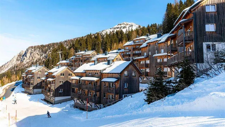 Eine Ansammlung von Holzhäusern in einer verschneiten Berglandschaft mit Skipiste und strahlendem Sonnenschein.