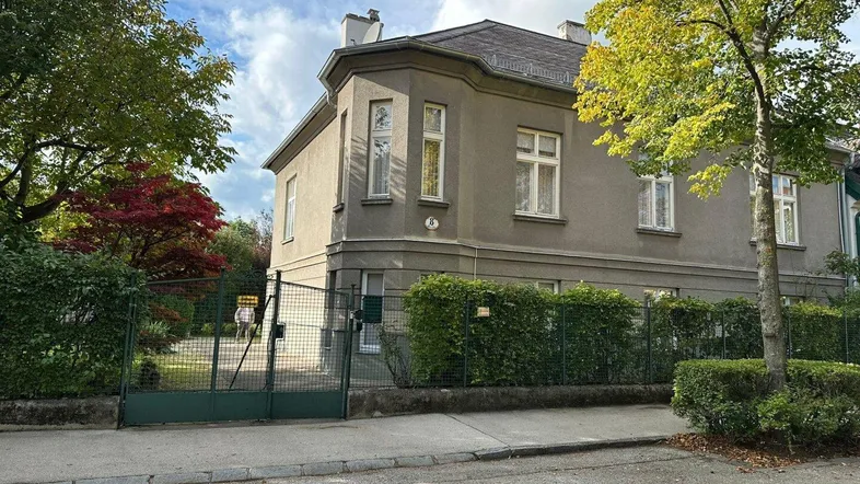 Klassisches Mehrfamilienhaus mit grauer Fassade und gepflegtem Vorgarten, umgeben von Bäumen und Hecken.