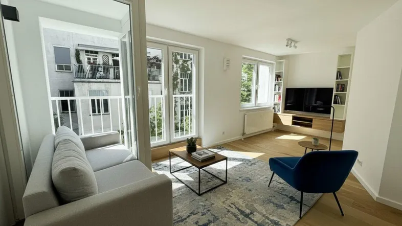Helles, modernes Wohnzimmer mit Holzboden, Zugang zum Balkon und integrierten Regalen.