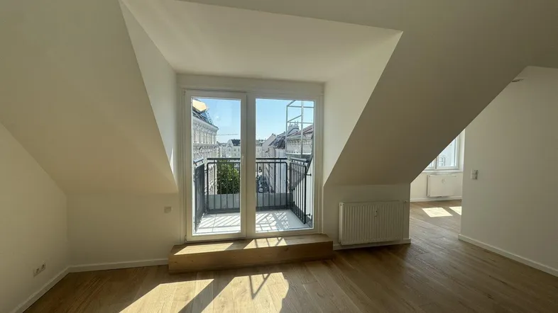 Heller Raum im Dachgeschoss mit Holzboden und Zugang zu einem kleinen Balkon mit Stadtblick.