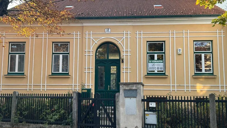 Gelbe Fassade eines historischen Gebäudes mit grüner Eingangstür und einem Verkaufsschild im Fenster.