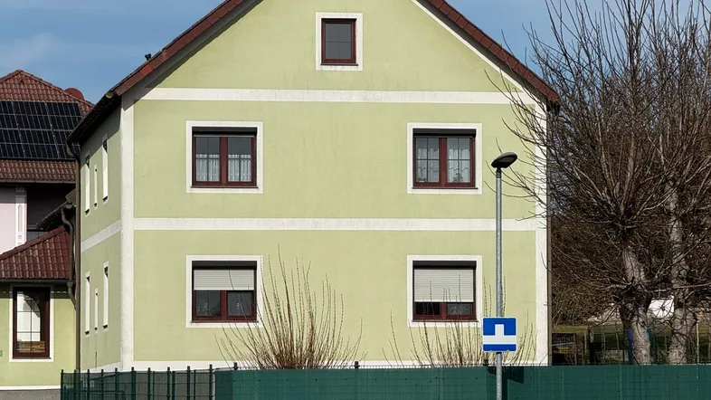 Vorderansicht eines Wohnhauses mit hellgrüner Fassade, rotem Ziegeldach und einem grünen Zaun.