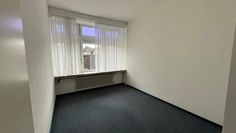 Kleiner, heller Raum mit Fenster, Heizkörper und dunklem gemustertem Teppichboden, ideal als Büro.