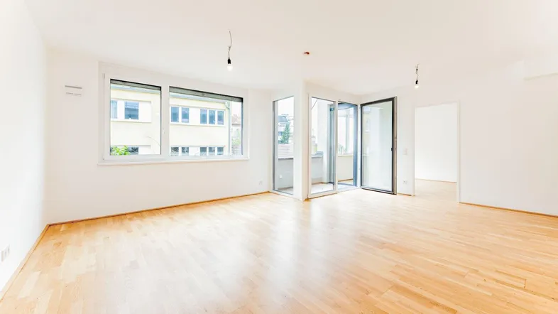 Helles, leeres Wohnzimmer mit Parkettboden und Zugang zu einem kleinen Balkon.