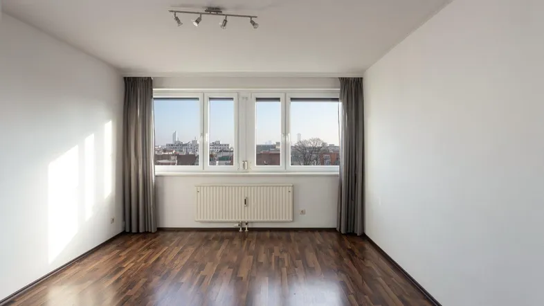 Helles Zimmer mit dunklem Parkettboden, weißen Wänden und Blick auf die Stadt.