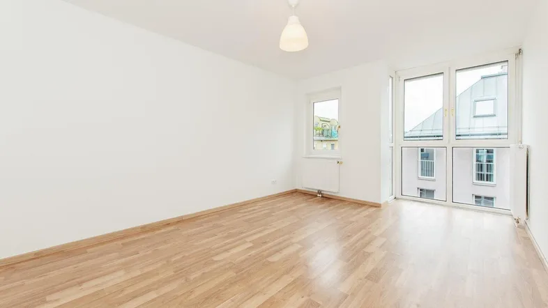 Helles, leeres Zimmer mit Laminatboden und großen Fenstern, die viel Tageslicht hereinlassen.