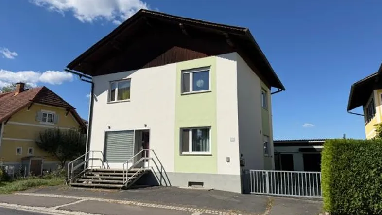 Freistehendes Einfamilienhaus mit weißer und hellgrüner Fassade unter blauem Himmel und Parkplätzen.