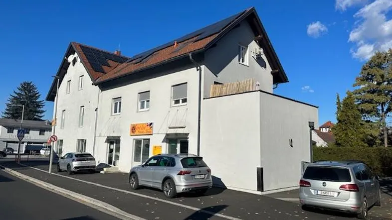 Weißes Gebäude mit Satteldach, Solarpaneelen und geparkten Autos auf der Straße.