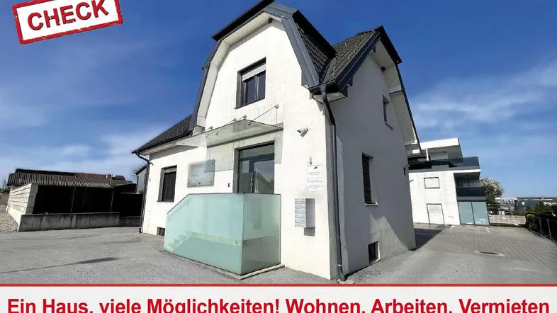 Weißes Gebäude mit dunklem Dach und Glasvordach, umgeben von einem gepflasterten Bereich.