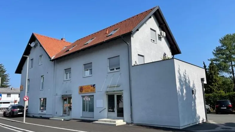 Gebäudeansicht mit hellgrauer Fassade, rotem Ziegeldach und Geschäftsflächen im Erdgeschoss.