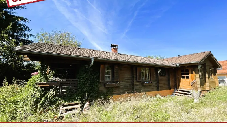 Rustikales Holzhaus mit braunem Dach und verwildertem Gartenbereich unter blauem Himmel.
