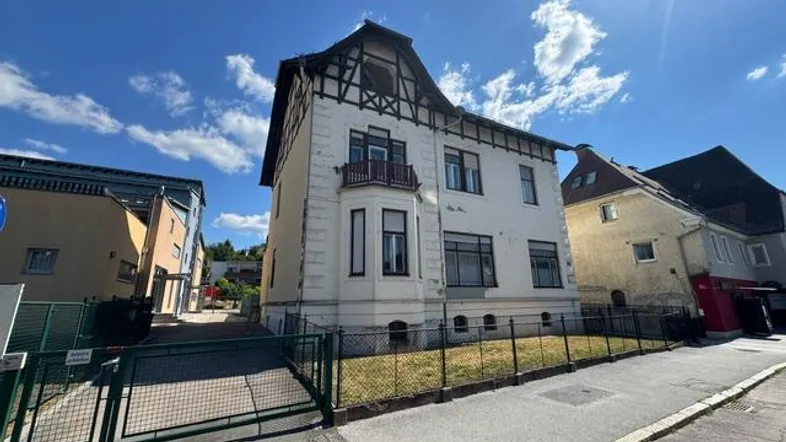 Historisches Gebäude mit markanter Fassade, Erker und traditionellem Dach, umgeben von einem Zaun und einer Straße.