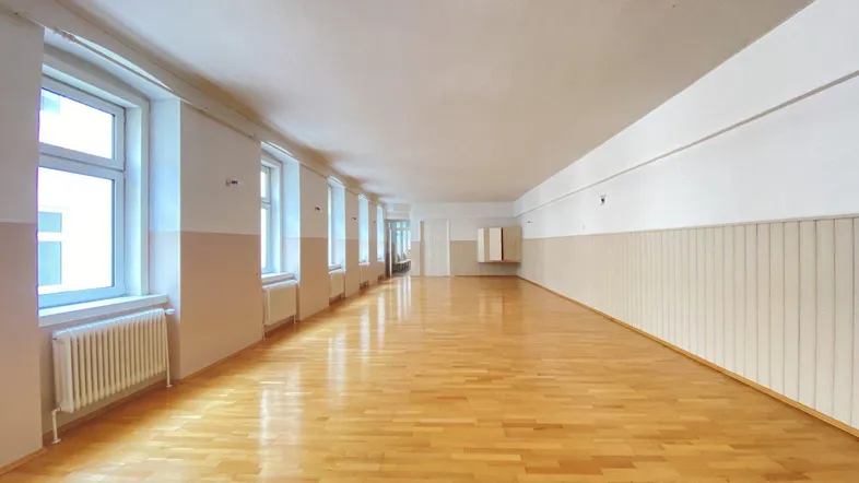 Langer, leerer Raum mit hellem Holzfußboden und vielen Fenstern, ideal für Büros oder Schulungsräume.