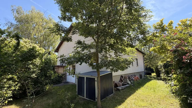 Seitenansicht des Hauses mit Garten, Rasenfläche und einem kleinen Geräteschuppen unter einem Baum.
