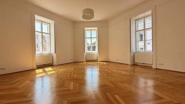 Helles Wohnzimmer mit Fischgrätparkett, hohen Decken und großen Fenstern, durch die Sonnenlicht fällt.