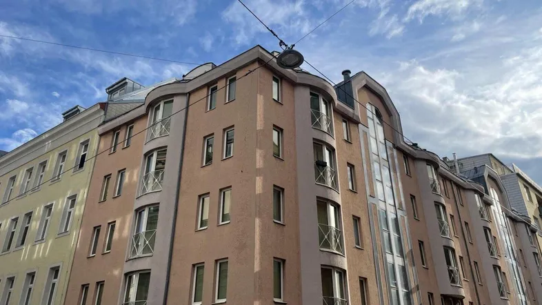 Gebäude an einer Straßenecke mit geschwungener Fassade und mehreren kleinen Balkonen unter bewölktem Himmel.