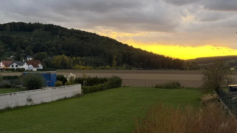 Sonnenuntergang über einem weiten Feld mit bewaldeten Hügeln und Häusern im Hintergrund.