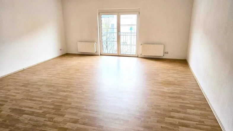Helles, leeres Wohnzimmer mit Laminatboden und Zugang zu einem französischen Balkon.