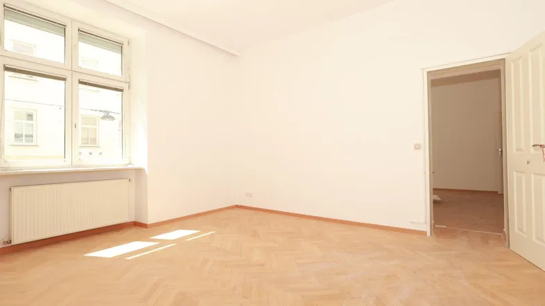 Geräumiges Zimmer mit hellem Fischgrätparkett, weißen Wänden und einem Fenster mit Blick nach draußen.