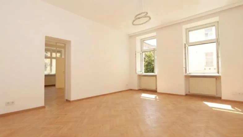 Helles Zimmer mit Fischgrätparkett, weißen Wänden und zwei Fenstern, das viel Tageslicht hereinlässt.