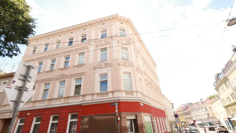 Mehrstöckiges Gebäude mit hellbeiger Fassade und roten Akzenten im Erdgeschoss an einer Straßenecke.
