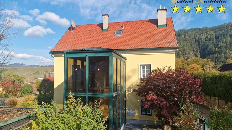 Gelbes Haus mit verglastem Wintergarten und rotem Ziegeldach vor bewaldeter Bergkulisse.