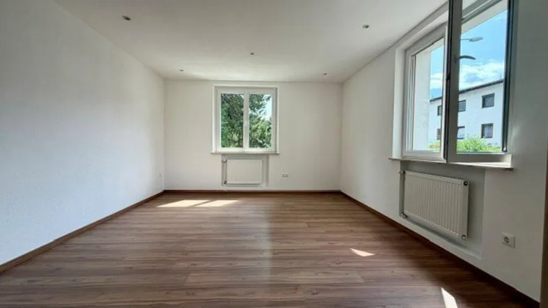 Helles, leeres Wohnzimmer mit Laminatboden und großen Fenstern, die viel Tageslicht hereinlassen.