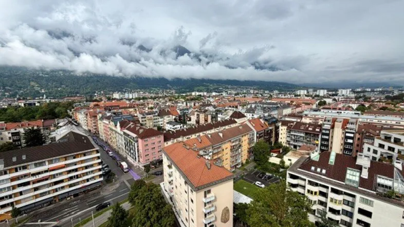 Panoramablick über eine Stadt mit Wohngebäuden und Bergen im Hintergrund unter bewölktem Himmel.