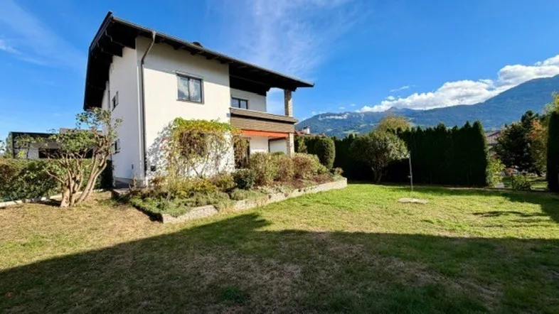 Weißes Einfamilienhaus mit großem, gepflegtem Garten und Bergblick unter strahlend blauem Himmel.