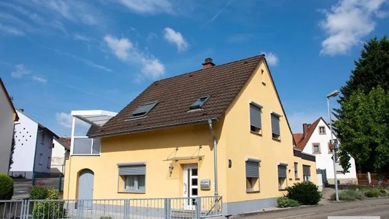 Gelbes Einfamilienhaus mit Satteldach, Rollläden und kleinem Vorgarten unter blauem Himmel.