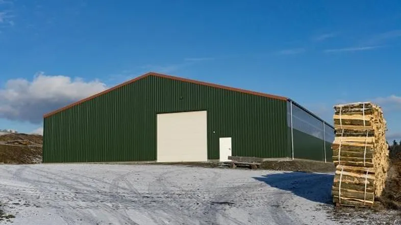 Große grüne Lagerhalle mit weißem Tor auf schneebedecktem Gelände unter blauem Himmel.