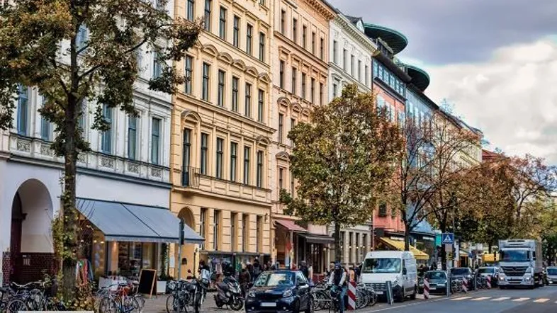 Belebte Straße mit historischen Gebäuden, Bäumen und parkenden Fahrzeugen in einer städtischen Umgebung.