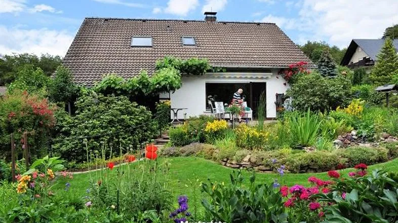 Haus mit gepflegtem Garten, blühenden Blumen und einer Terrasse mit Sitzgelegenheiten im Grünen.