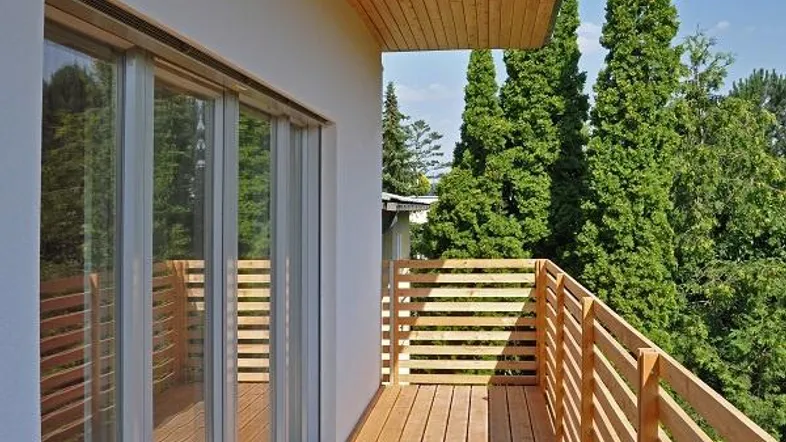 Heller Balkon mit Holzboden und Holzgeländer, umgeben von hohen Bäumen und viel Grün.