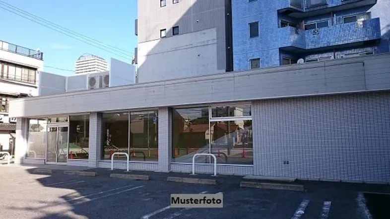 Gebäude mit großen Schaufenstern im Erdgeschoss und darüber liegenden Wohneinheiten, umgeben von einem Parkplatz.