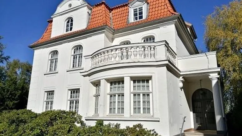 Elegante weiße Villa mit rotem Ziegeldach und einem geschwungenen Balkon, umgeben von grünen Büschen.