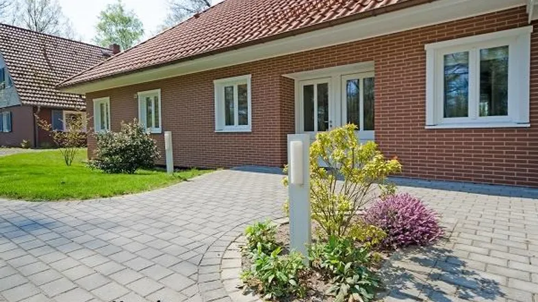 Ziegelhaus mit rotem Ziegeldach, weißen Fenstern und gepflastertem Vorplatz mit kleiner Bepflanzung.