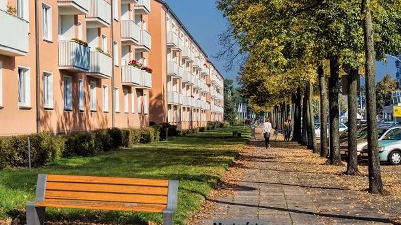 Wohngebäude mit Balkonen, umgeben von Bäumen und einem Gehweg mit Bank in einer grünen Umgebung.