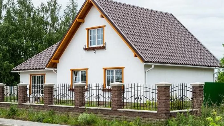 Weißes Einfamilienhaus mit braunem Ziegeldach, Holzfenstern und einem dekorativen Zaun im Vordergrund.