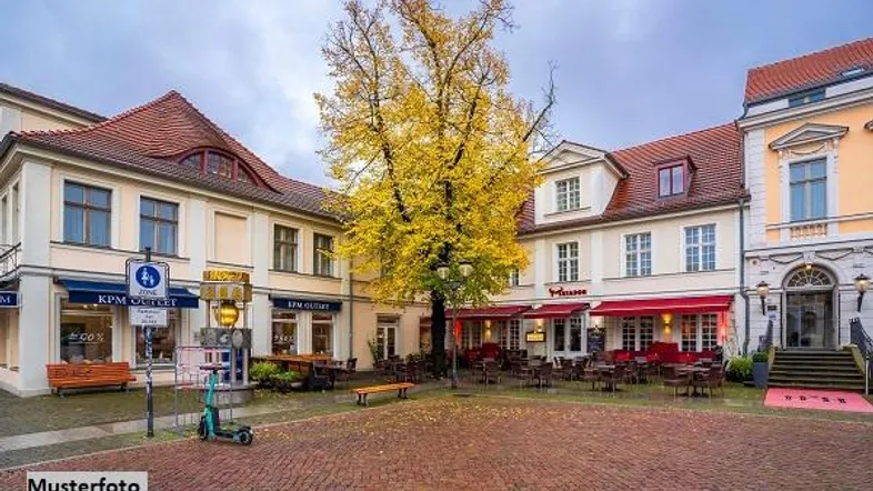 Belebter Stadtplatz mit historischen Gebäuden, Außengastronomie und einem großen Baum mit Herbstlaub.