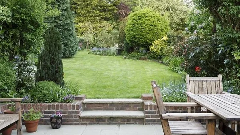 Gepflegter Garten mit großer Rasenfläche, altem Baumbestand und einer Terrasse mit Holzmöbeln.
