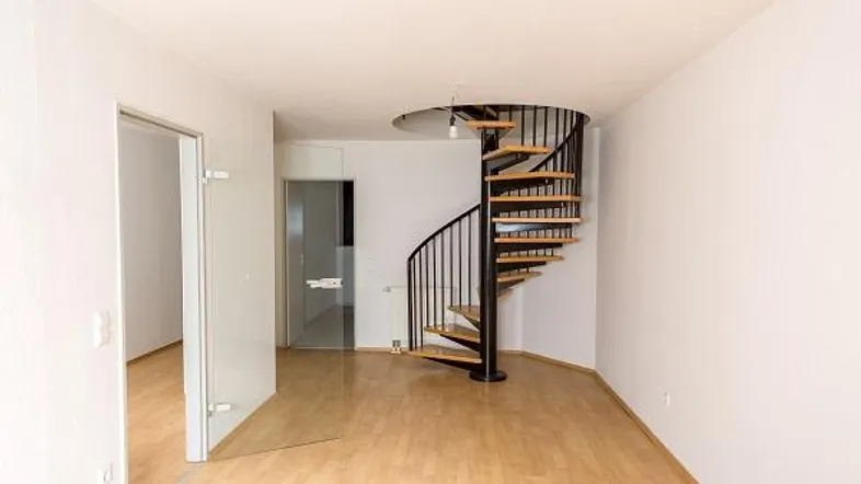 Heller, leerer Raum mit Holzfußboden und einer eleganten schwarzen Wendeltreppe, die in die obere Etage führt.