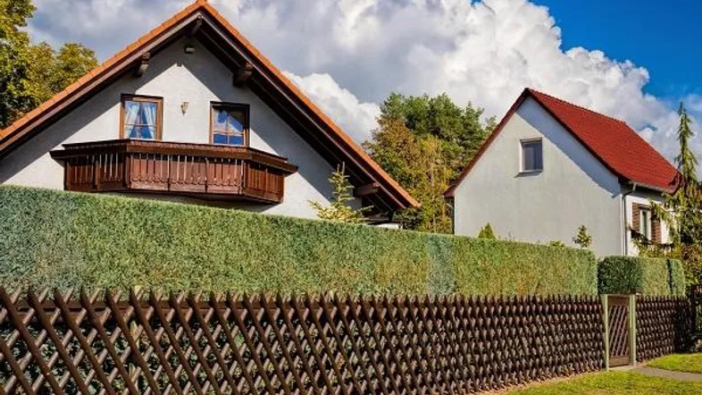 Zwei gepflegte Häuser mit roten Ziegeldächern, umgeben von einem Holzzaun und einer hohen grünen Hecke.